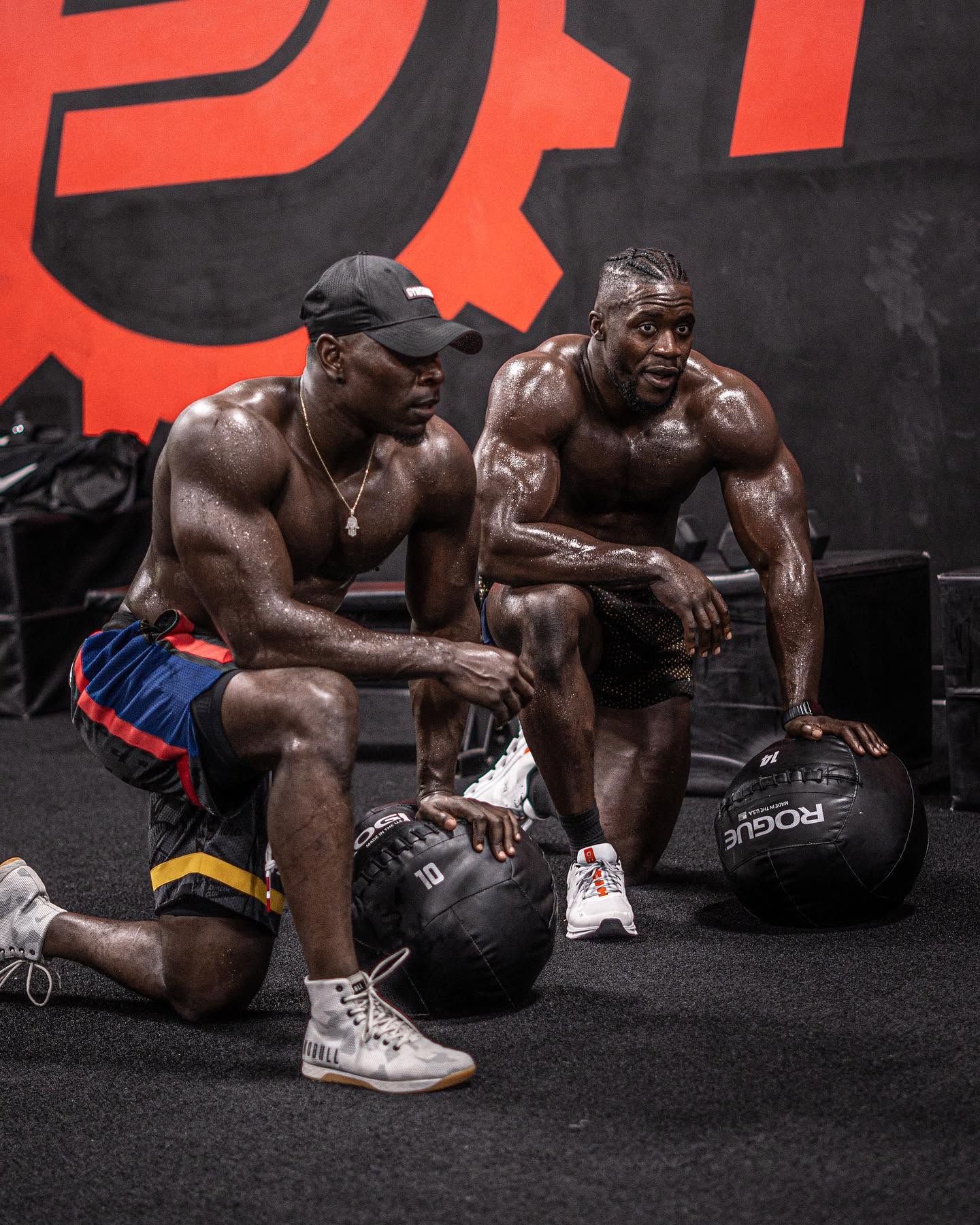 Two muscular athletes kneel with kettlebells in a gym, sweat-covered and resting after a workout.
