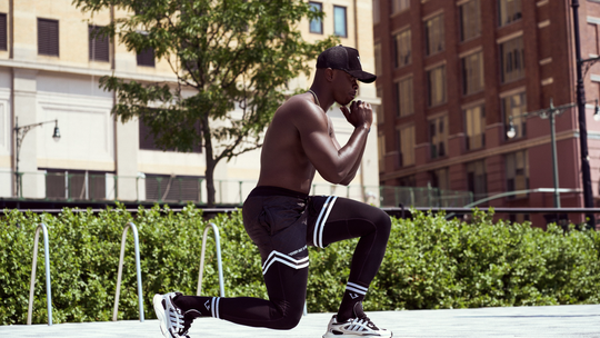 Athlete lunging outdoors on a city sidewalk, wearing black workout gear and a cap.