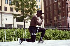 Person stretching in a lunge outdoors beside a hedge and city buildings