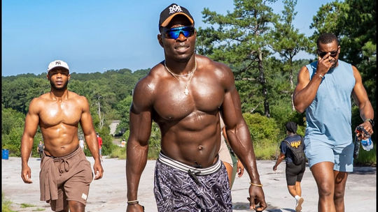 Three men walking on a sandy beach, with the center man shirtless and others in casual summer clothes.