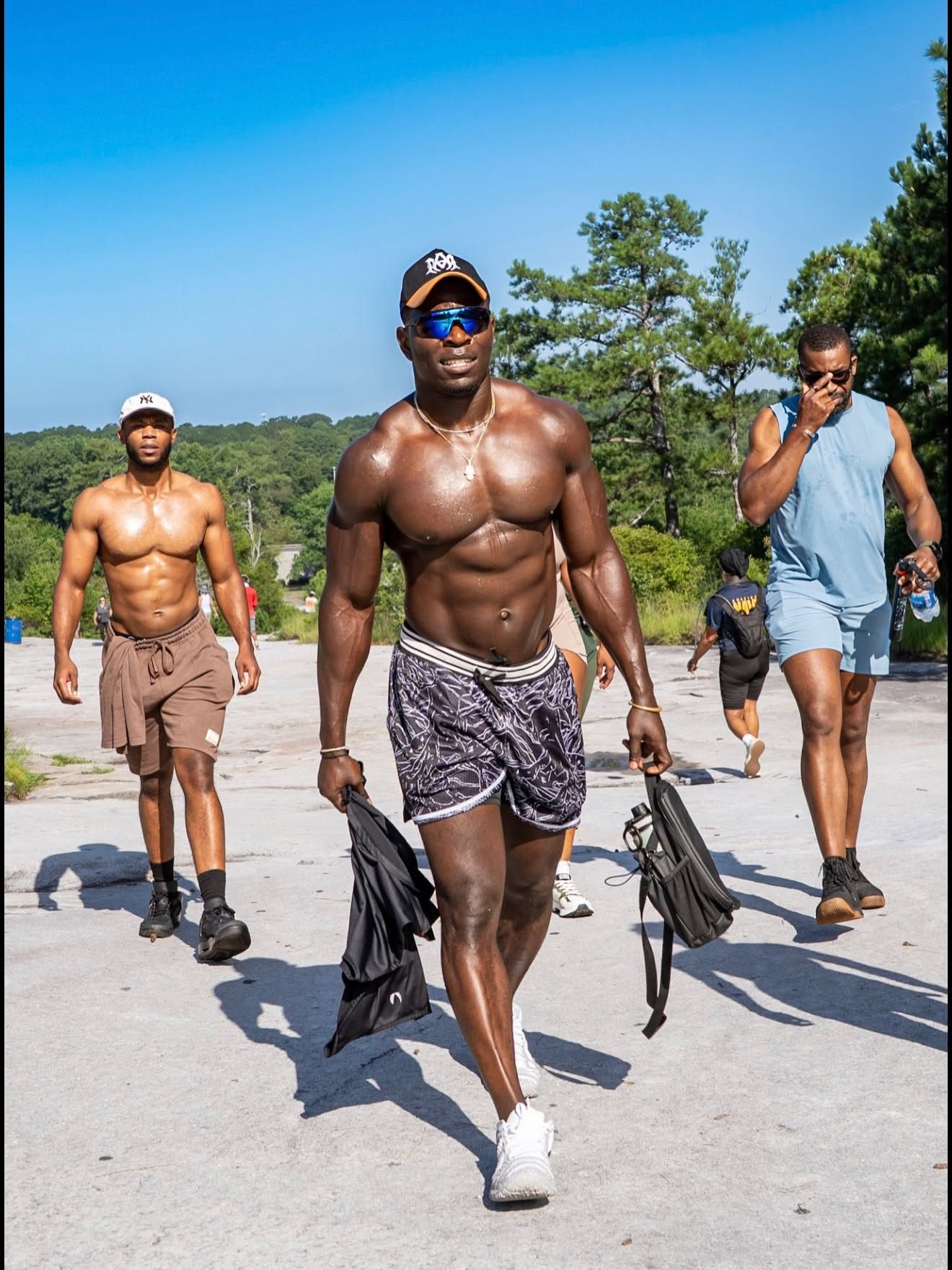 Three shirtless men walk on a sunny road carrying flippers, with palm trees and blue sky behind them.