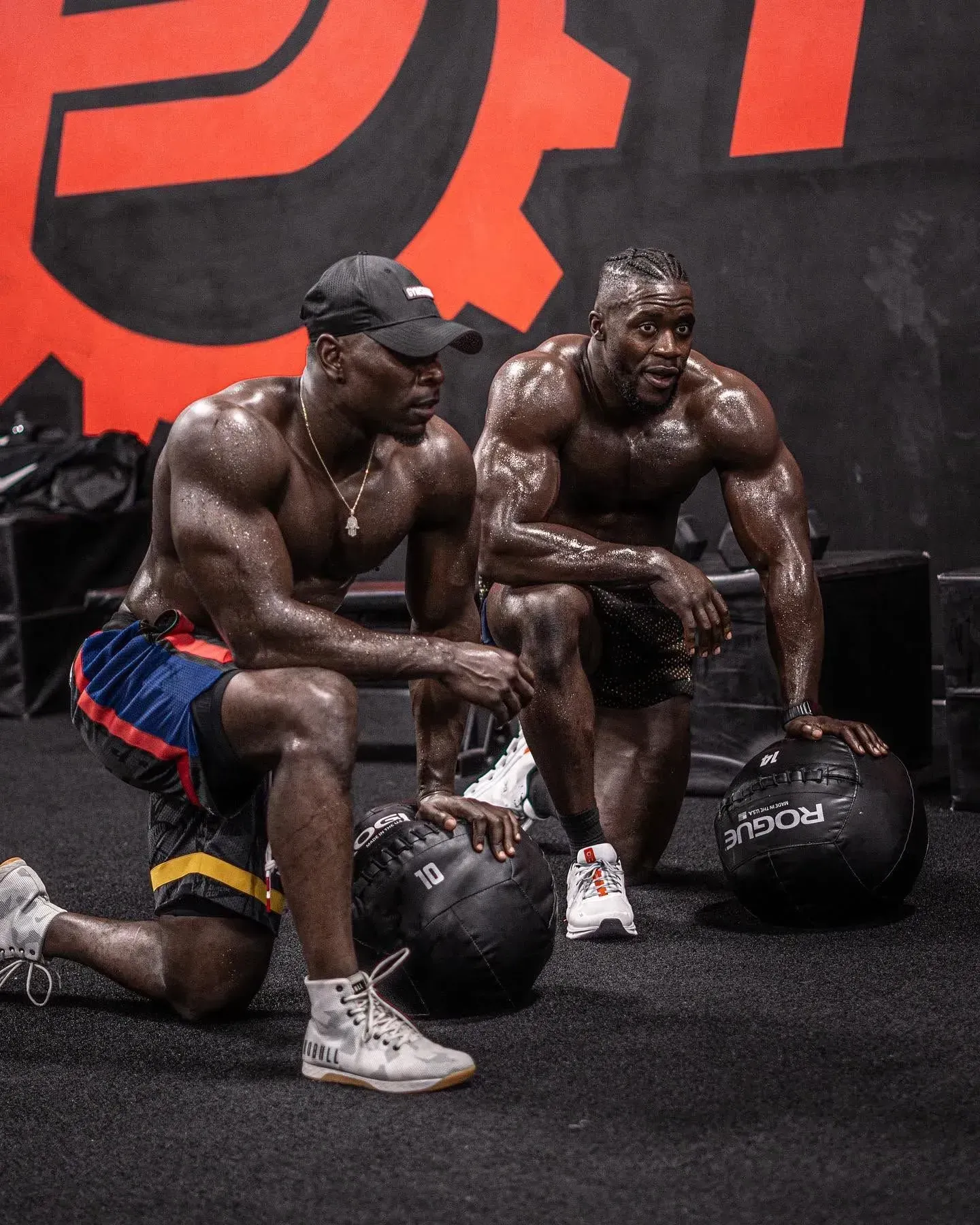 Two muscular athletes kneel with kettlebells in a gym, sweat-covered and resting after a workout.