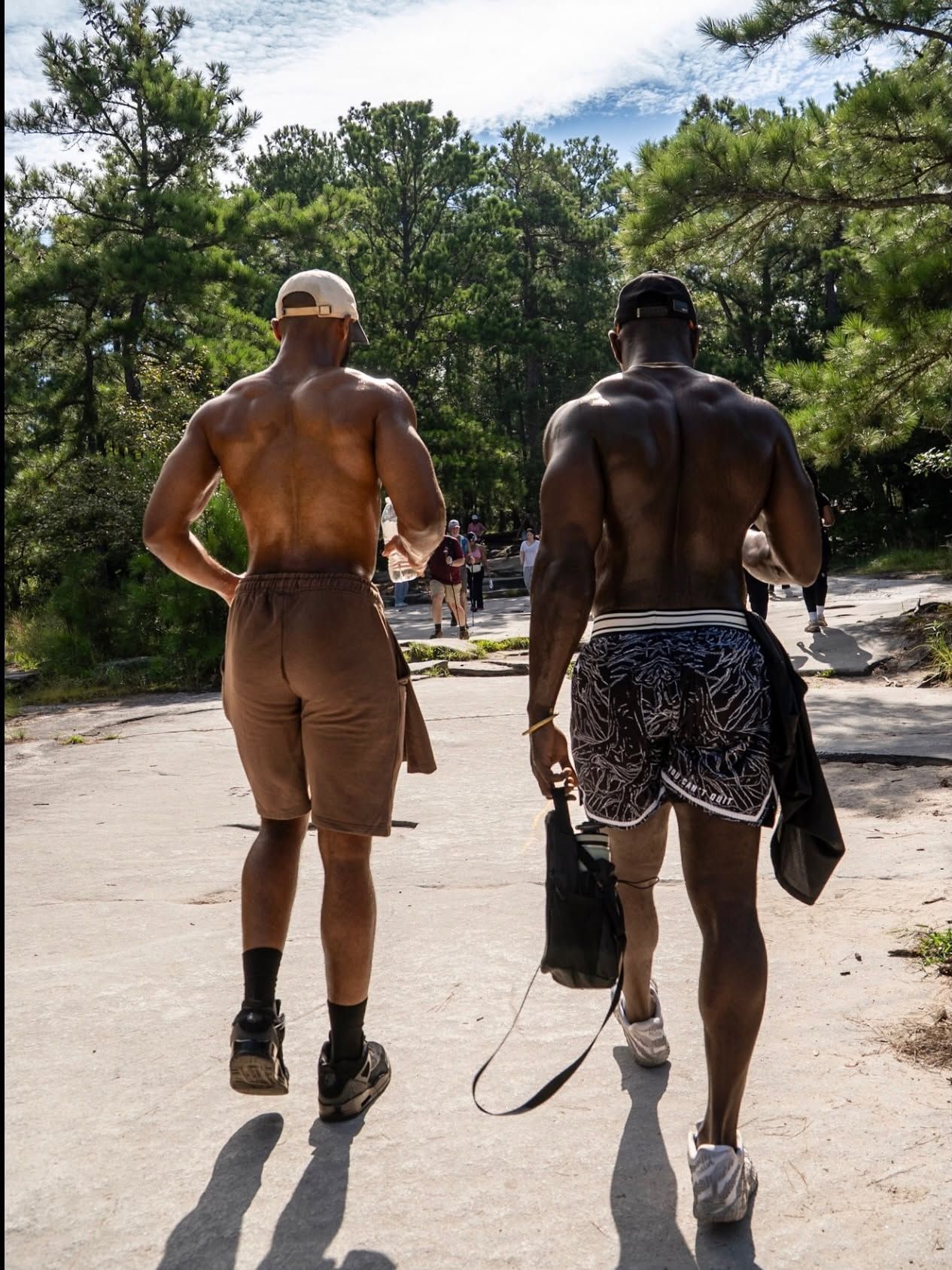 Two shirtless men walking on a sunny wooded trail, one carrying a towel and the other wearing patterned shorts.