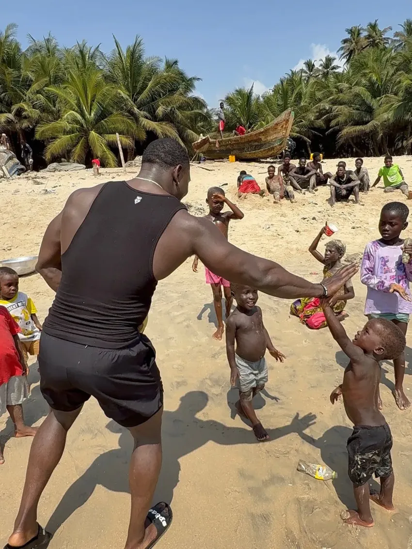 Man in black shorts dancing with children on a sandy beach, with palm trees and nets in the background