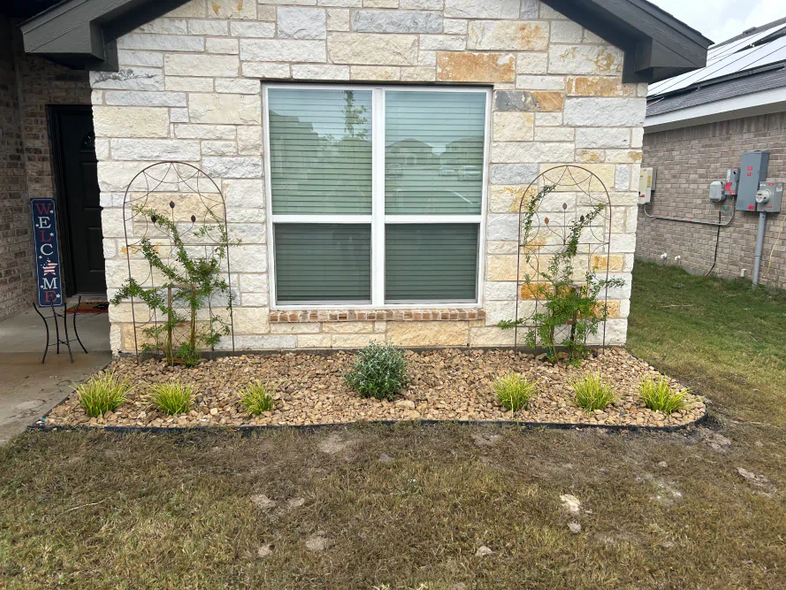 Front of a beige stone house with a centered window and small landscaped flower beds in the yard