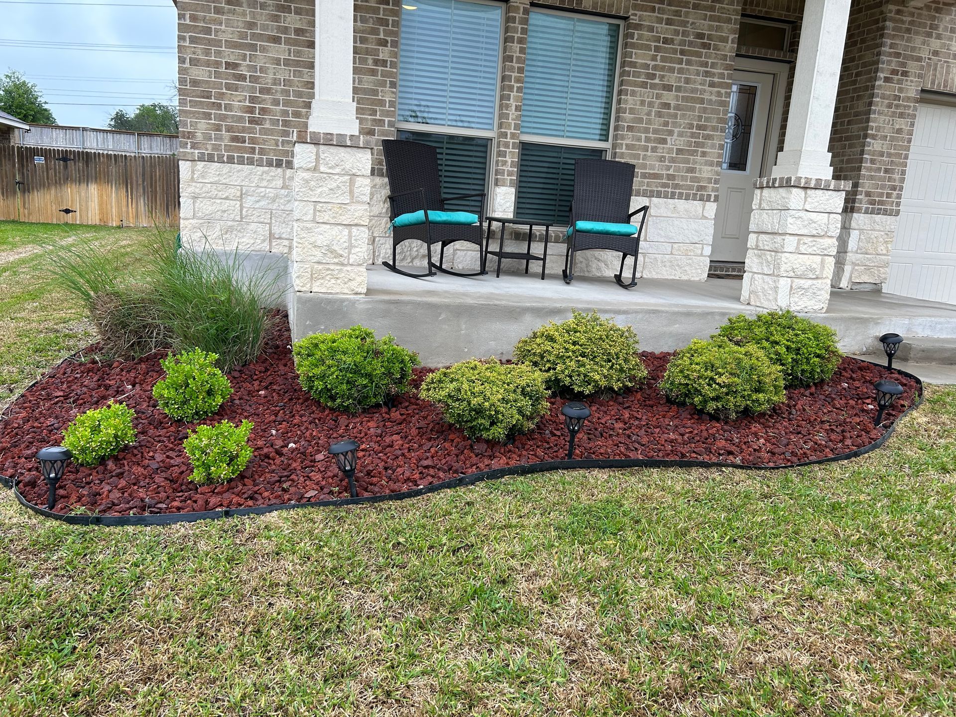 Front porch with two chairs and a curved landscaped flower bed of green shrubs and red mulch.