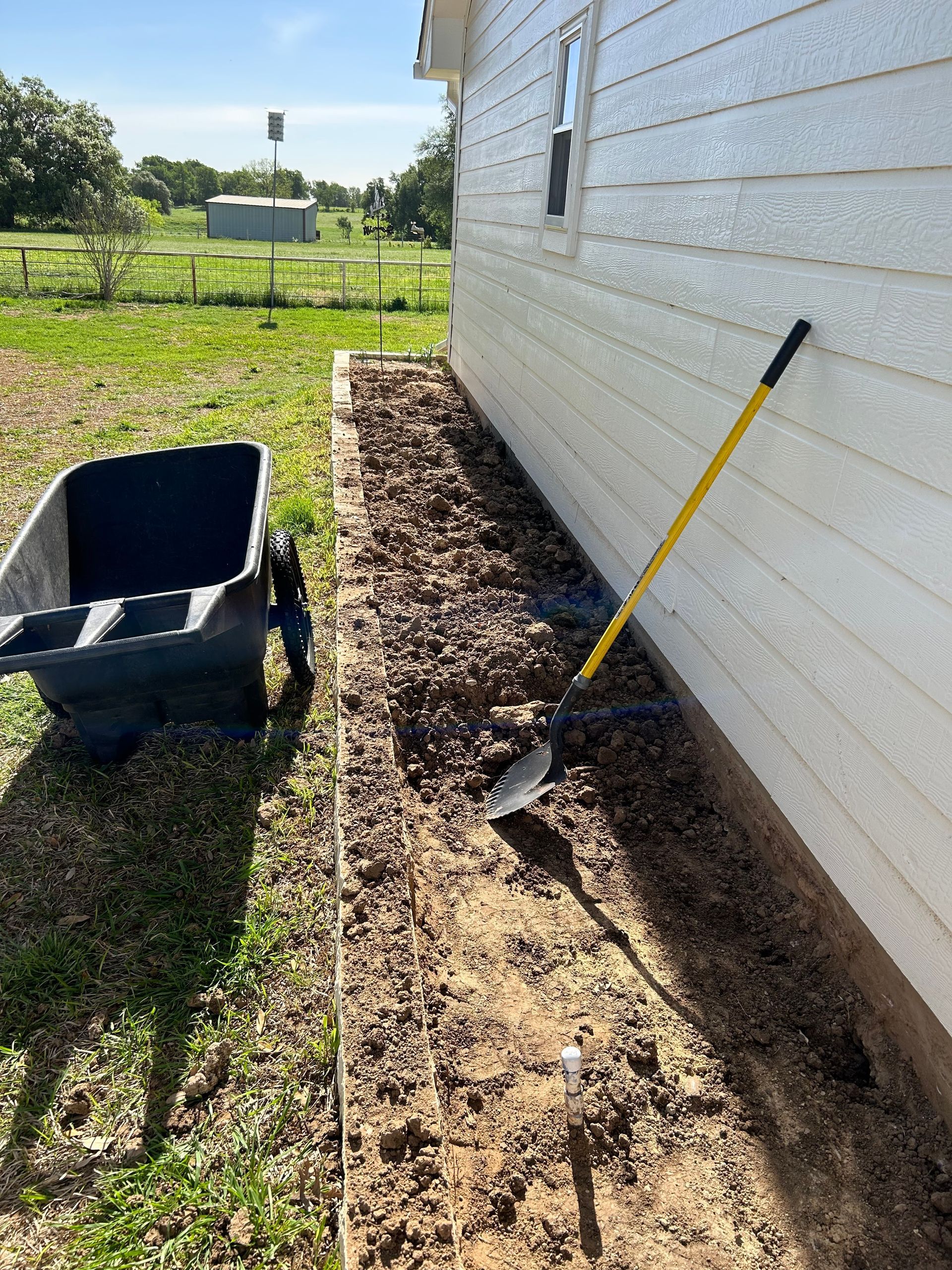 Gardening tools beside a house wall in a sunlit yard, with soil and mulch along the foundation