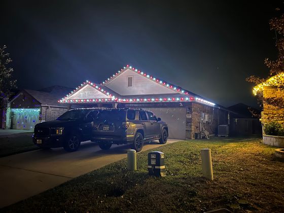 Nighttime house decorated with white and red Christmas lights, with two parked vehicles in the driveway.