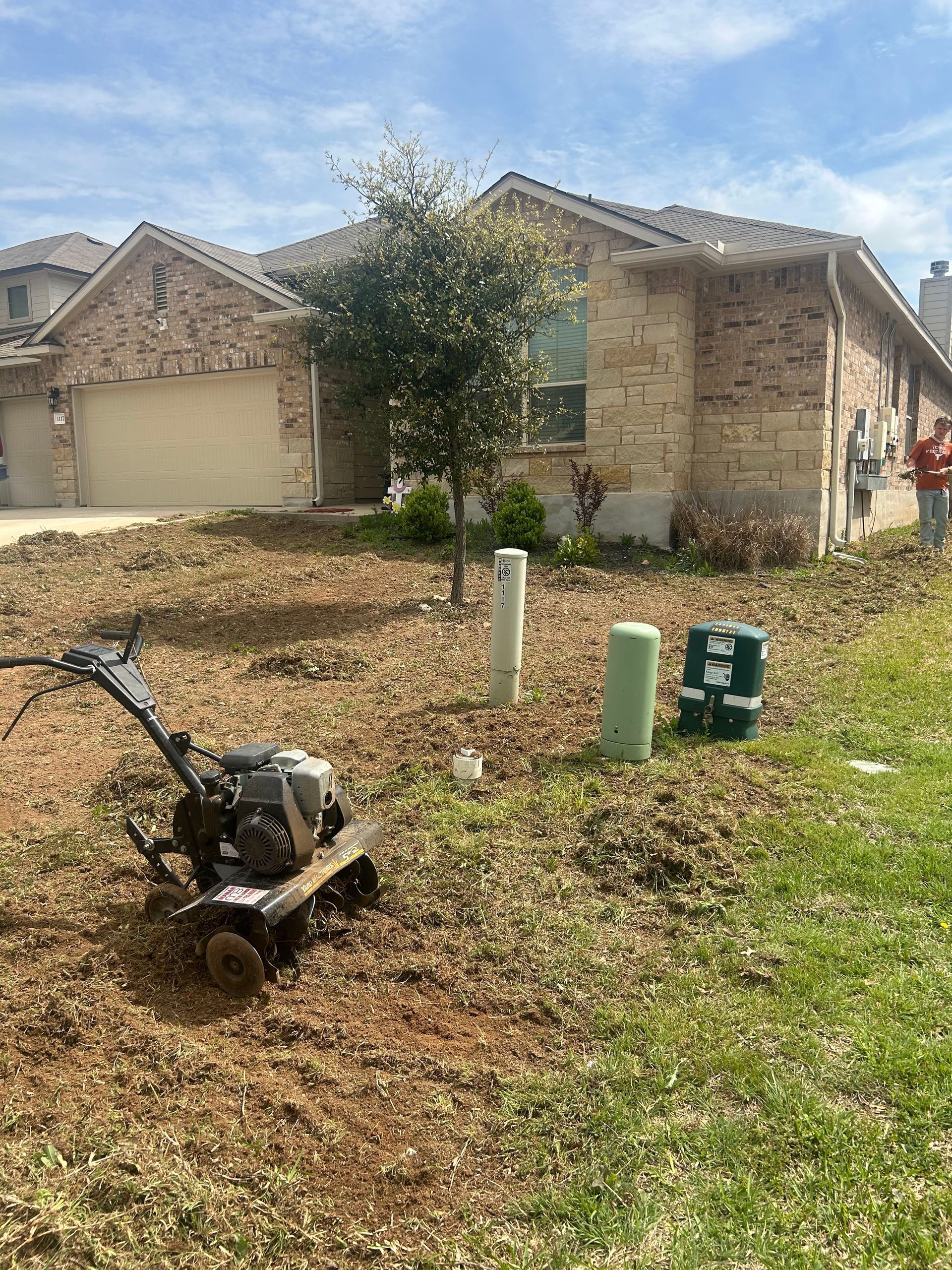 Front yard with a wheelbarrow, tree, utility boxes, and a stone house with a garage in the background