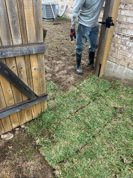 Person standing in a muddy yard beside an open wooden gate, holding a tool.