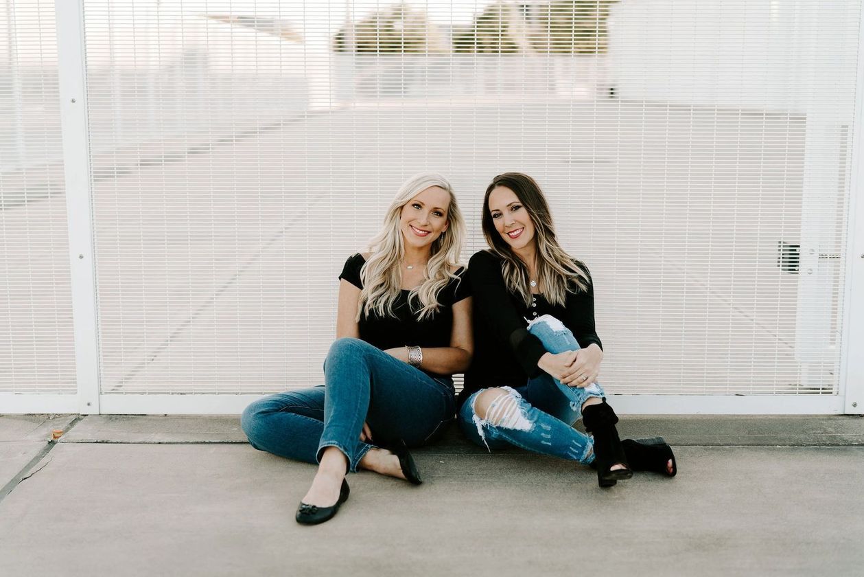 Two women sitting on the ground in front of a white fence, smiling at the camera.