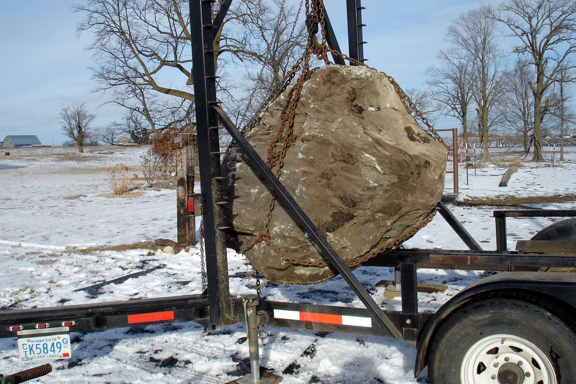 A large rock suspended by chains from a rig, on a trailer in a snowy field.