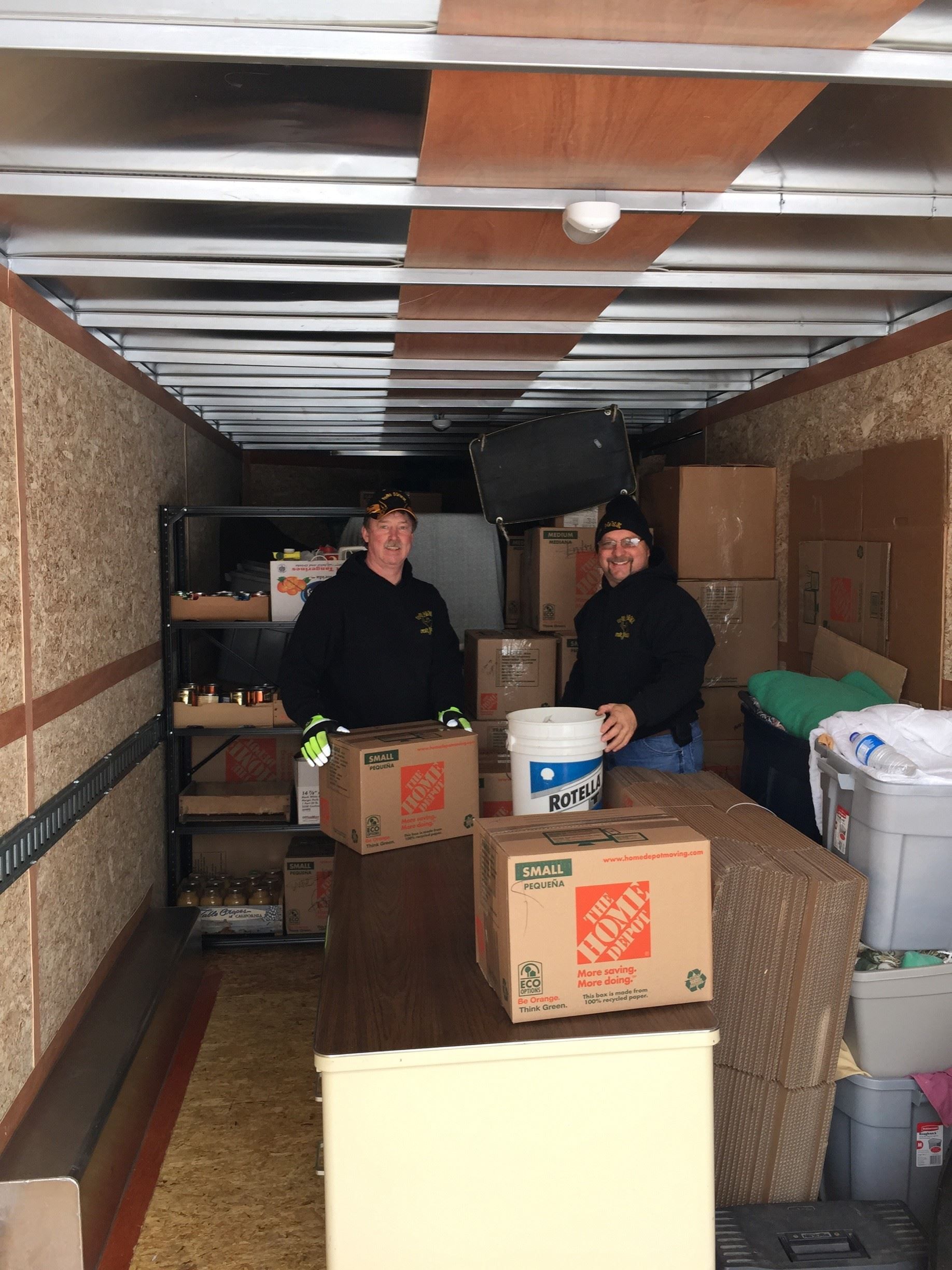 Two people in a moving truck, holding boxes. Boxes, furniture, and other items fill the truck.