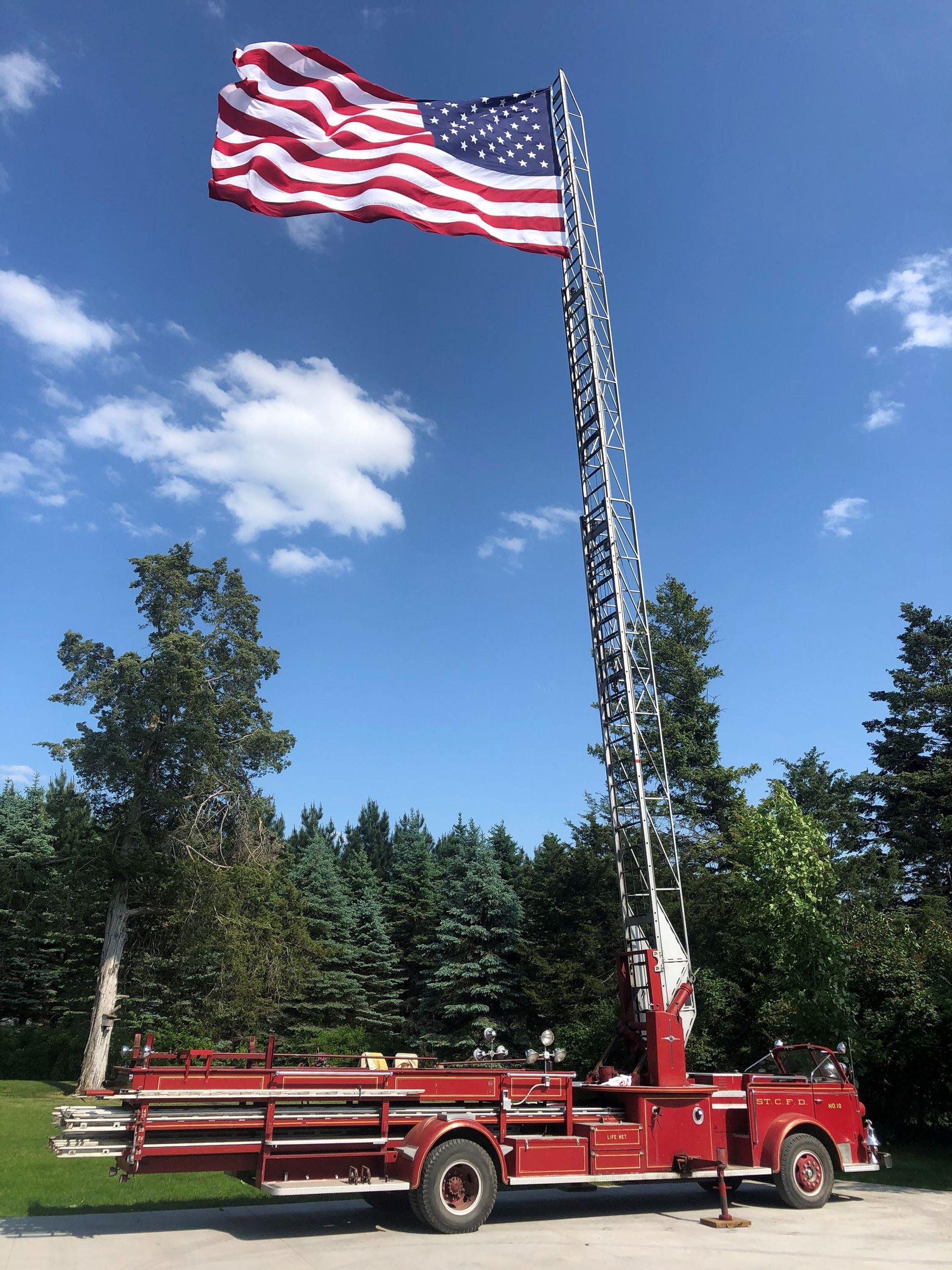 Fire truck with extended ladder holding a large American flag against a blue sky.