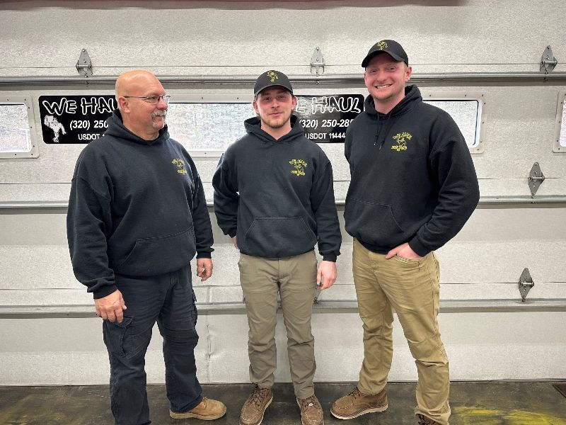 Three men in black hoodies stand in front of a garage door, smiling. One has a hand in his pocket.