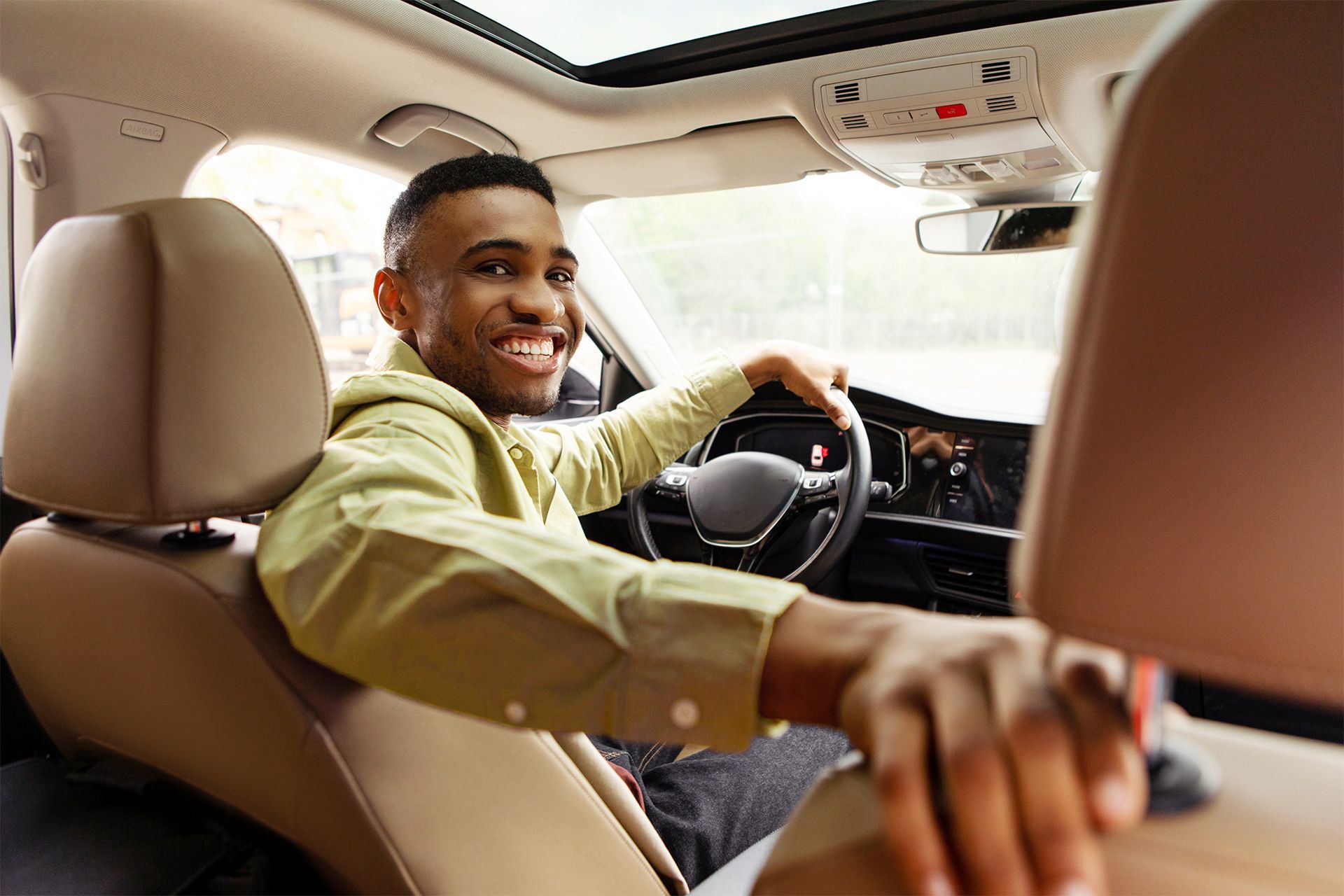 Smiling person driving a car, looking back. Inside of a vehicle, headrest and steering wheel visible.