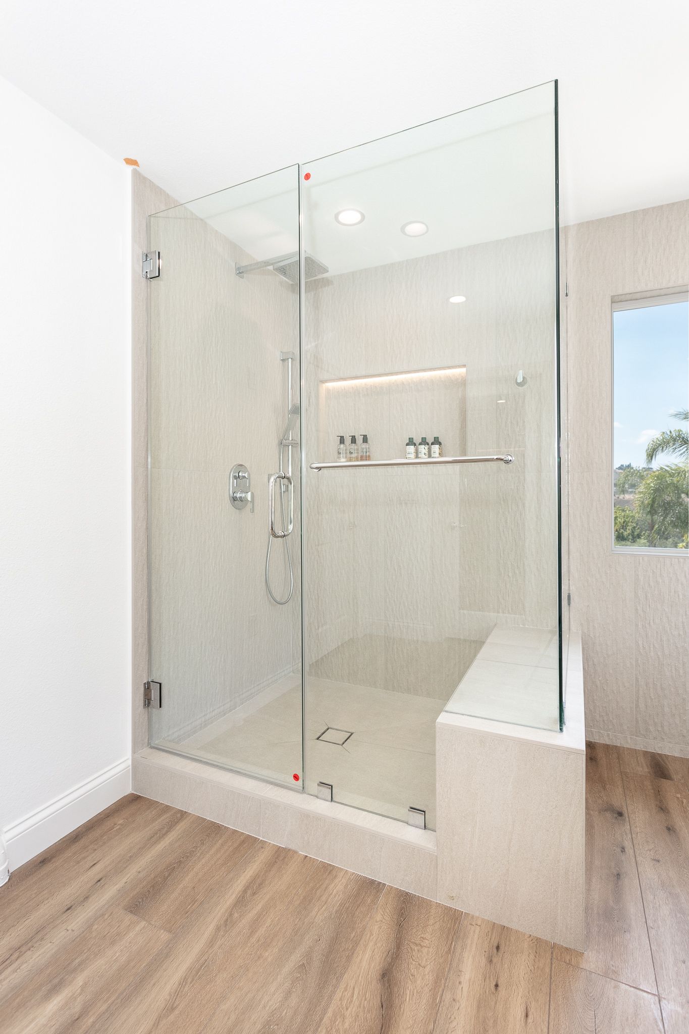 Glass shower with bench seat; tan walls, wood floor, chrome fixtures. Bright bathroom.