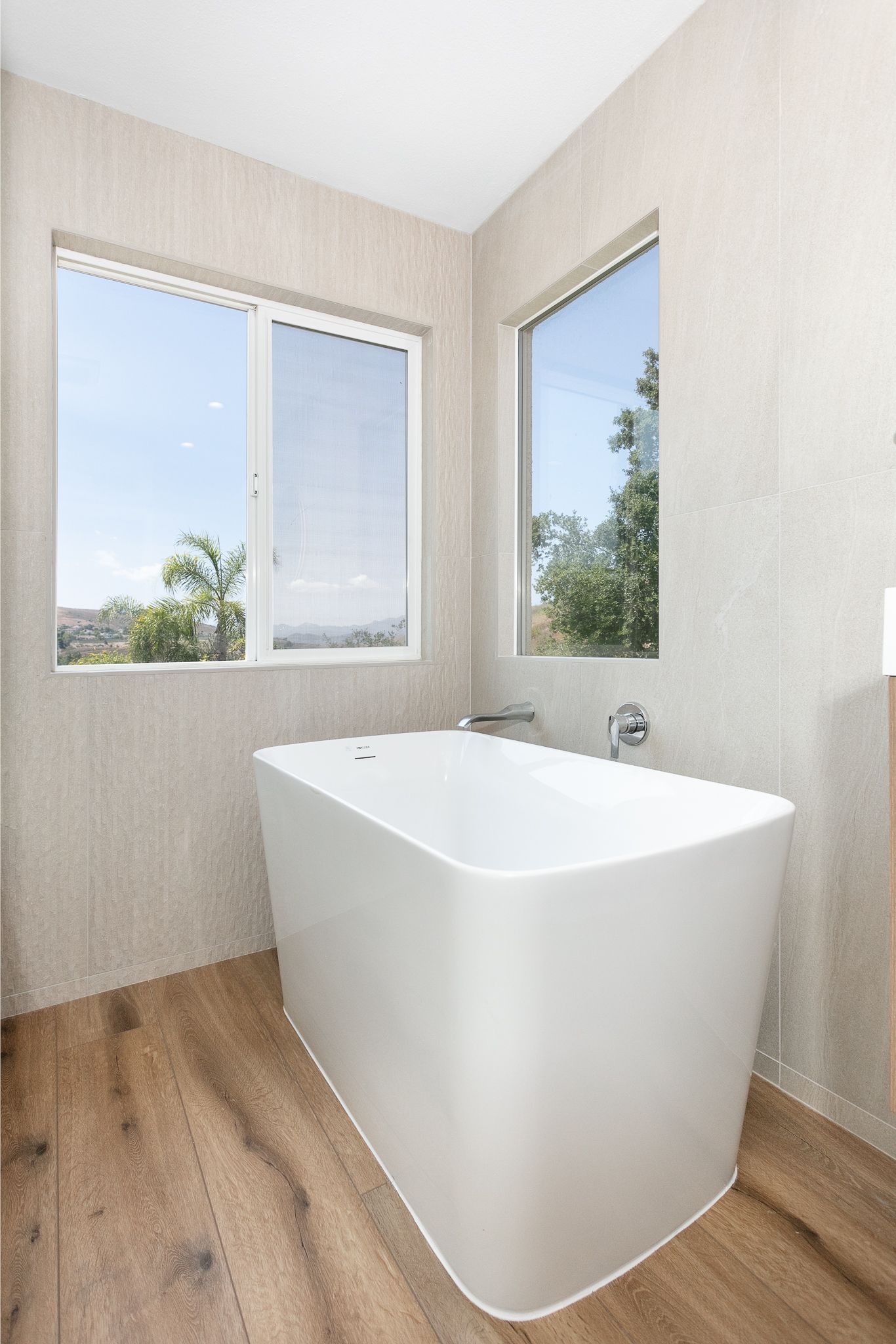 Modern white freestanding bathtub in a bathroom with windows and wood-look flooring.