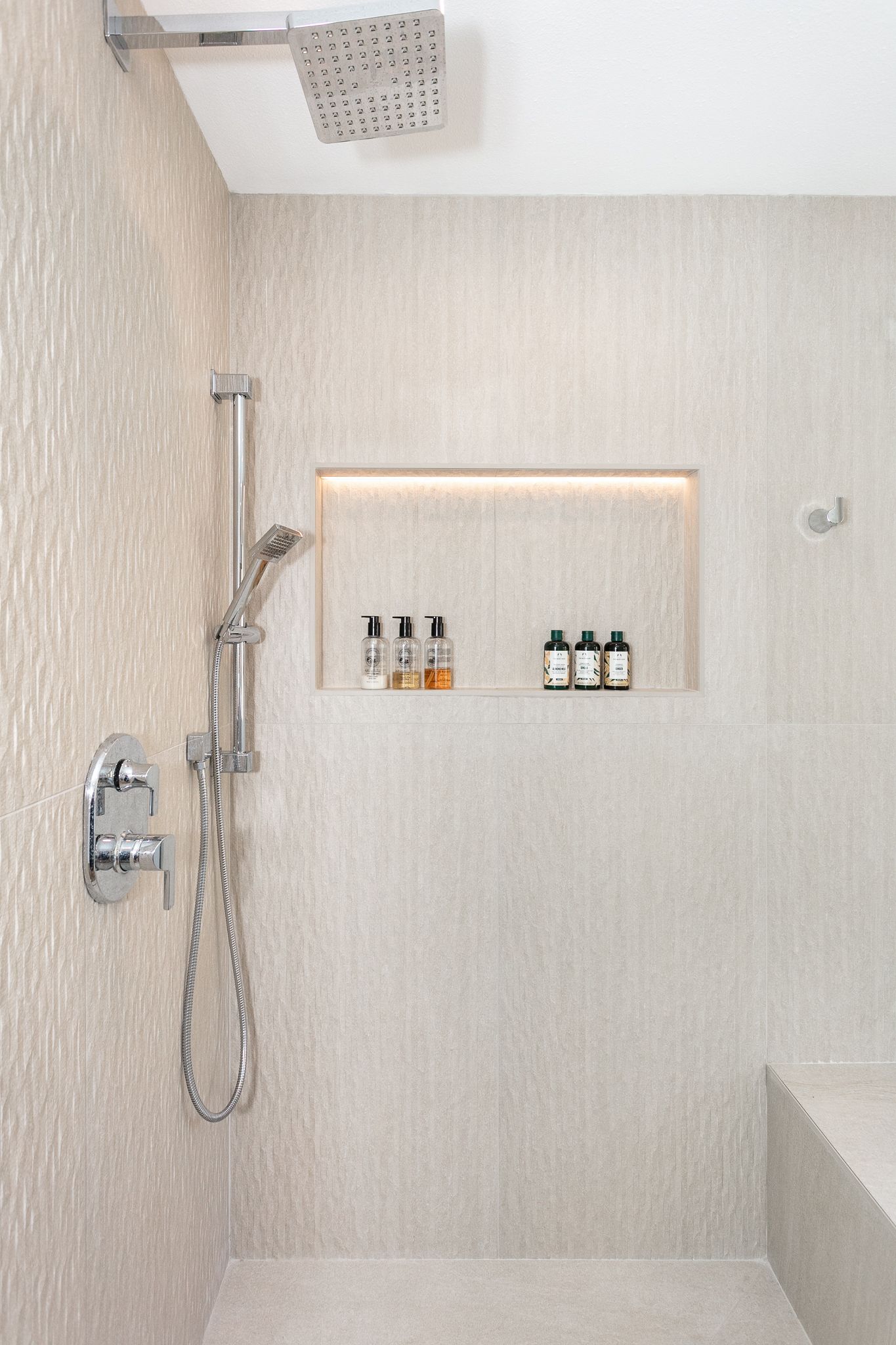 Shower with textured beige tile and a built-in shelf holding toiletries; features a rain shower head and handheld wand.