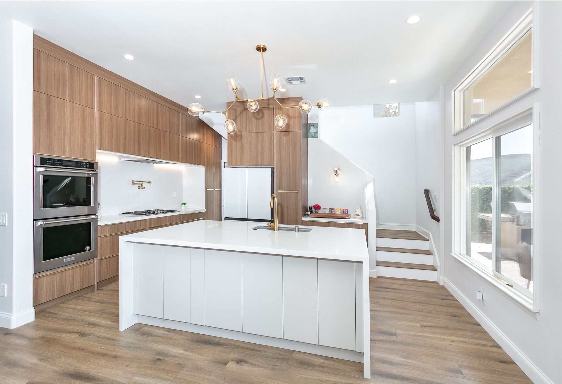 Modern kitchen with white island, wood cabinets, gold chandelier, and stairs to upper level.