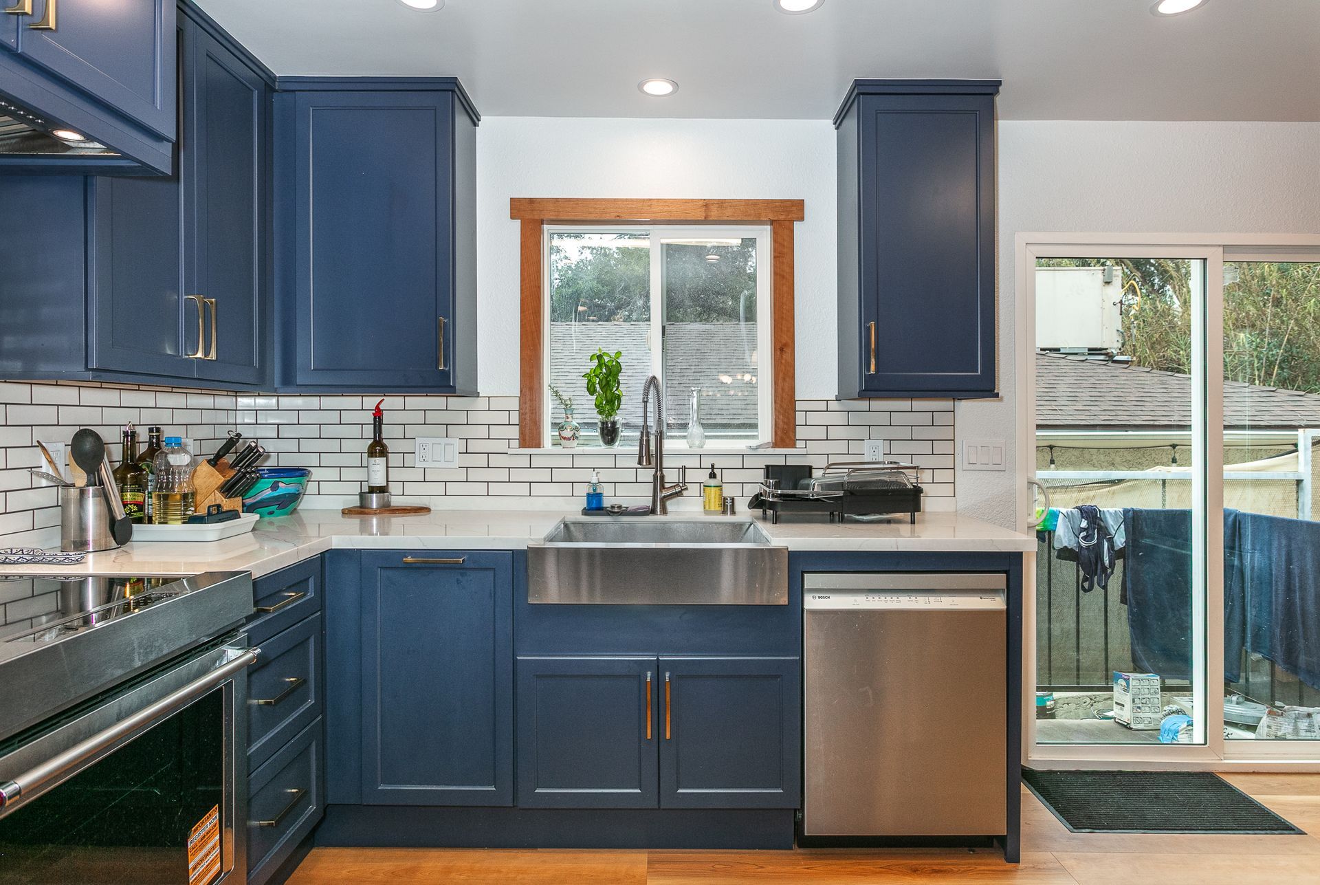 Blue kitchen cabinets with white backsplash and stainless steel sink. Sliding glass door and wood-framed window.