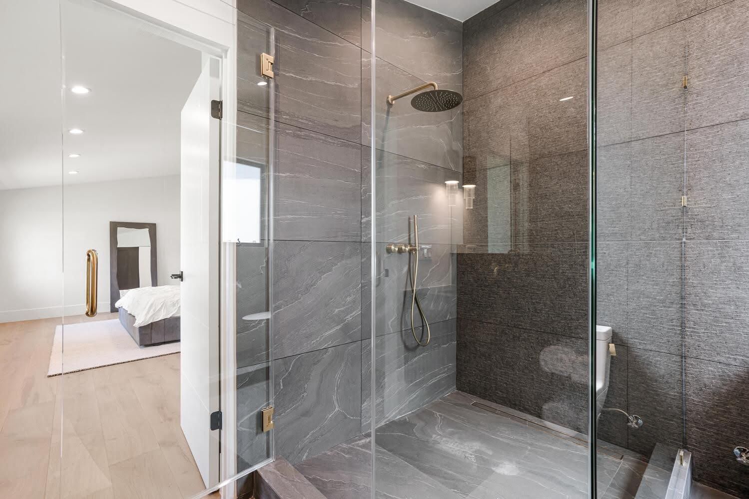 Modern bathroom with gray tile shower, glass door, and a glimpse of bedroom.