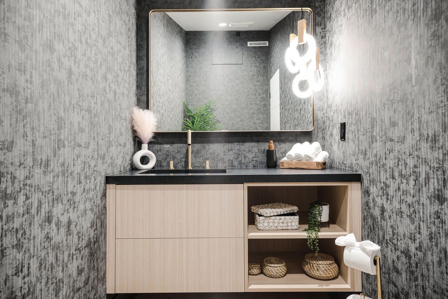 Bathroom with textured gray walls, light wood vanity, gold accents, and a decorative light fixture.