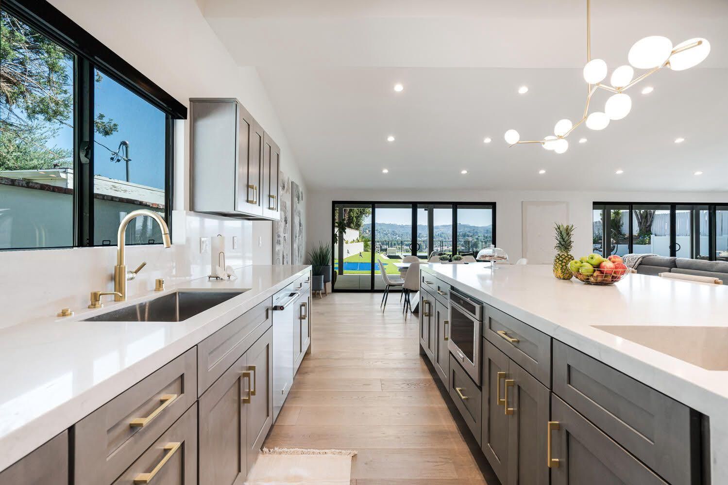 Modern kitchen with gray cabinets, gold hardware, and large windows overlooking a pool and scenic view.