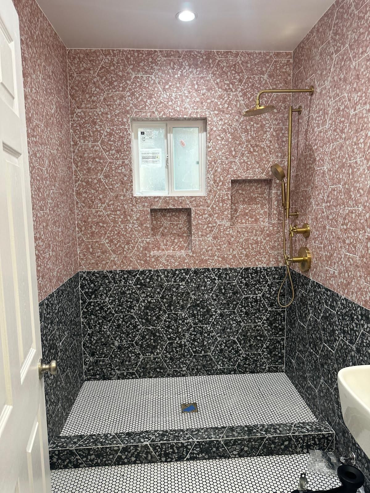 A bathroom with pink tile walls, black/white mosaic shower floor and black tile half wall. Gold shower fixtures.
