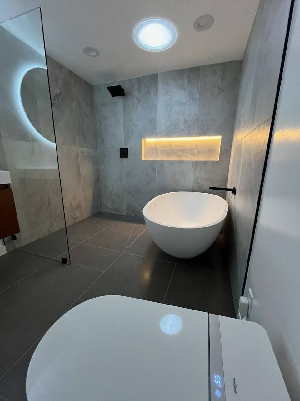Modern bathroom with a white tub, toilet, and gray tiled walls and floor.
