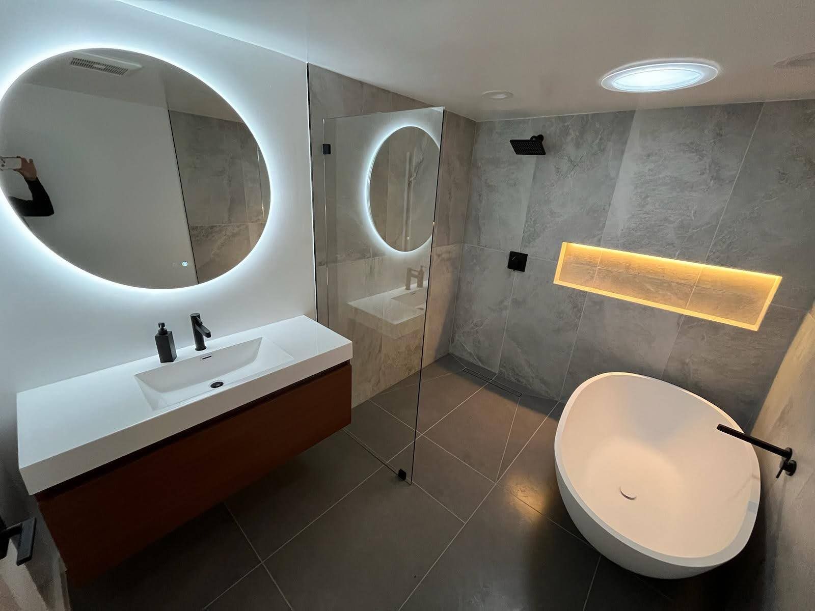Modern bathroom with floating vanity, round backlit mirrors, glass shower, and a freestanding tub.