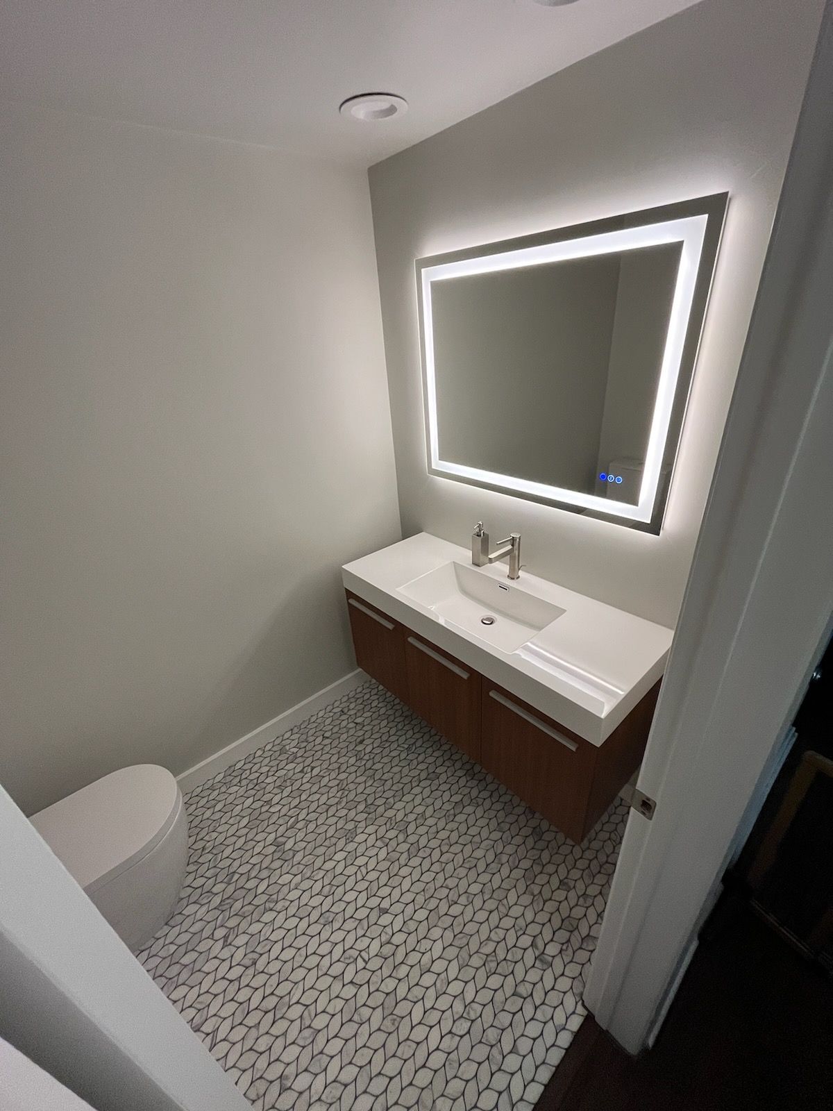 Small, modern bathroom with a floating vanity, lighted mirror, and white and gray tile floor.