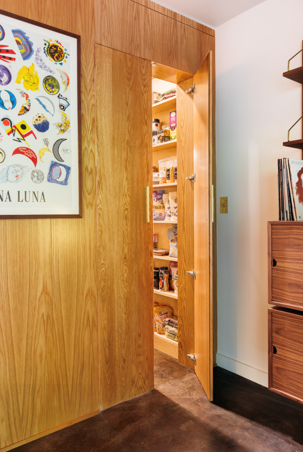 Wooden pantry door ajar, revealing shelves of food. Light wood paneling, art on wall, dark floor.