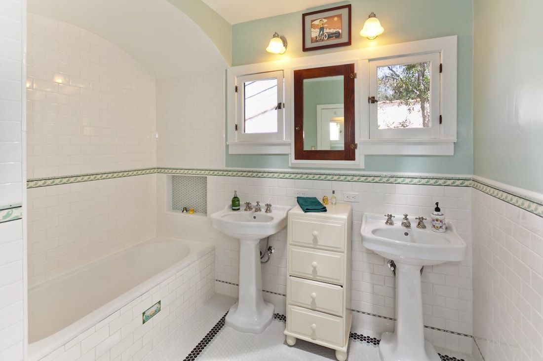 Bathroom with white tile, pedestal sinks, a bathtub, and a small dresser. Light blue walls and a dark wood mirror.