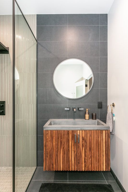 Guest bathroom is adorned with large dark grey tiles continuing on the wall behind a custom floating walnut vanity with a fluted design, complemented by a concrete sink and a black wall-mounted faucet. The shower enclosure separating the vanity from the shower area consists of a glass panel with a black frame to complement the black hardware and trim theme.
