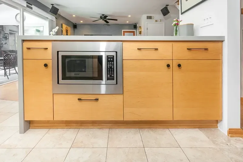 Kitchen cabinetry with built-in microwave, light wood finish, stainless steel accents, and beige tile flooring.