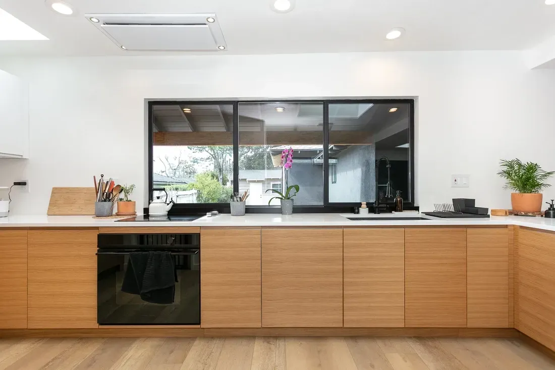 Modern kitchen with light wood cabinets, white countertops, large window, and black oven.