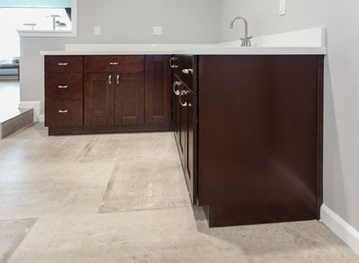 Dark wood L-shaped bar cabinets with white countertop, faucet, and drawers on light tile floor.