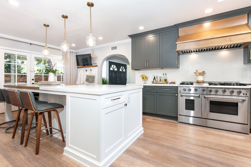 Modern kitchen: white island with seating, blue-gray cabinets, stainless steel appliances, light wood floors.
