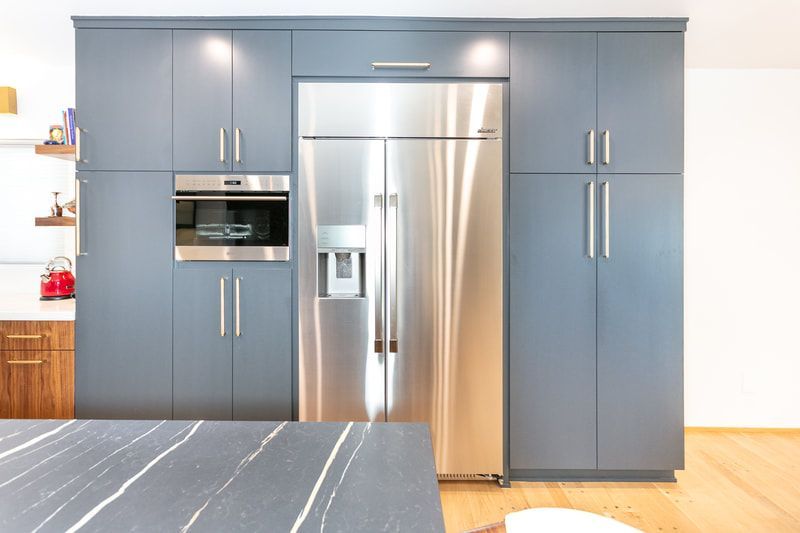 Blue kitchen cabinets surround a stainless steel refrigerator and built-in oven, with a dark countertop in the foreground.