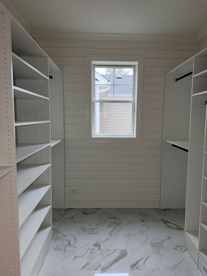 Narrow white walk-in closet with built-in shelves, marble-pattern floor, and a small window.