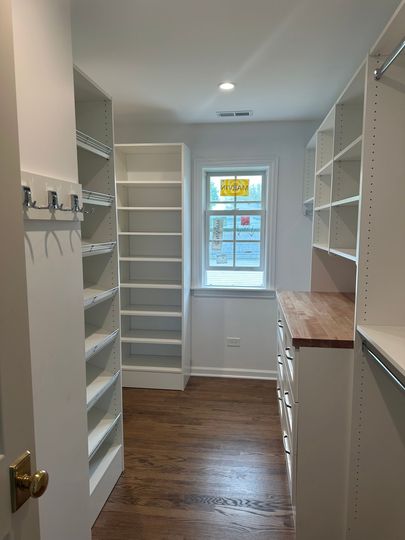 Narrow walk-in closet with white shelves, wood floor, and a window at the far end.