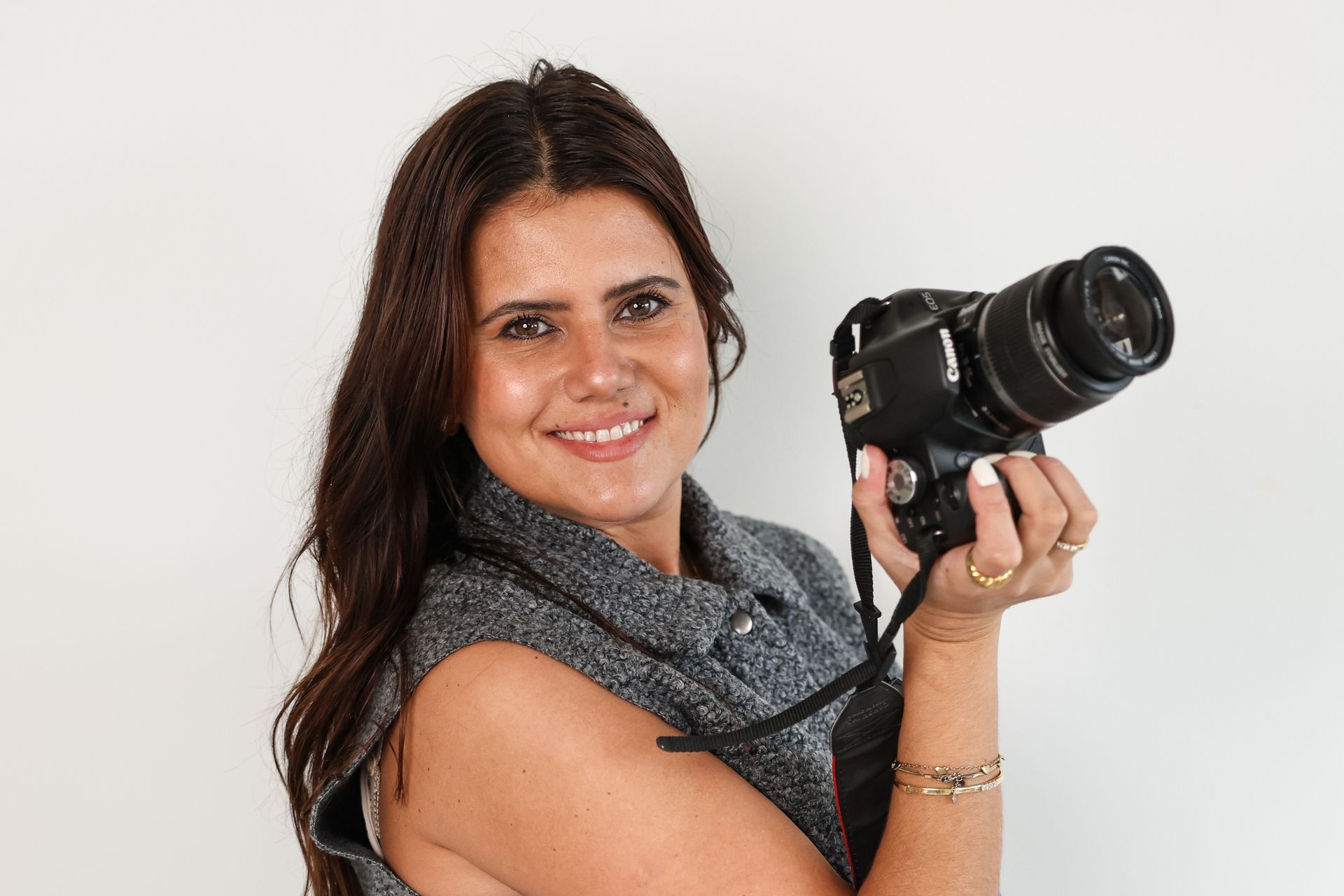 Een lachend persoon met lang donker haar houdt een digitale camera vast, gekleed in een grijs shirt, tegen een witte achtergrond.