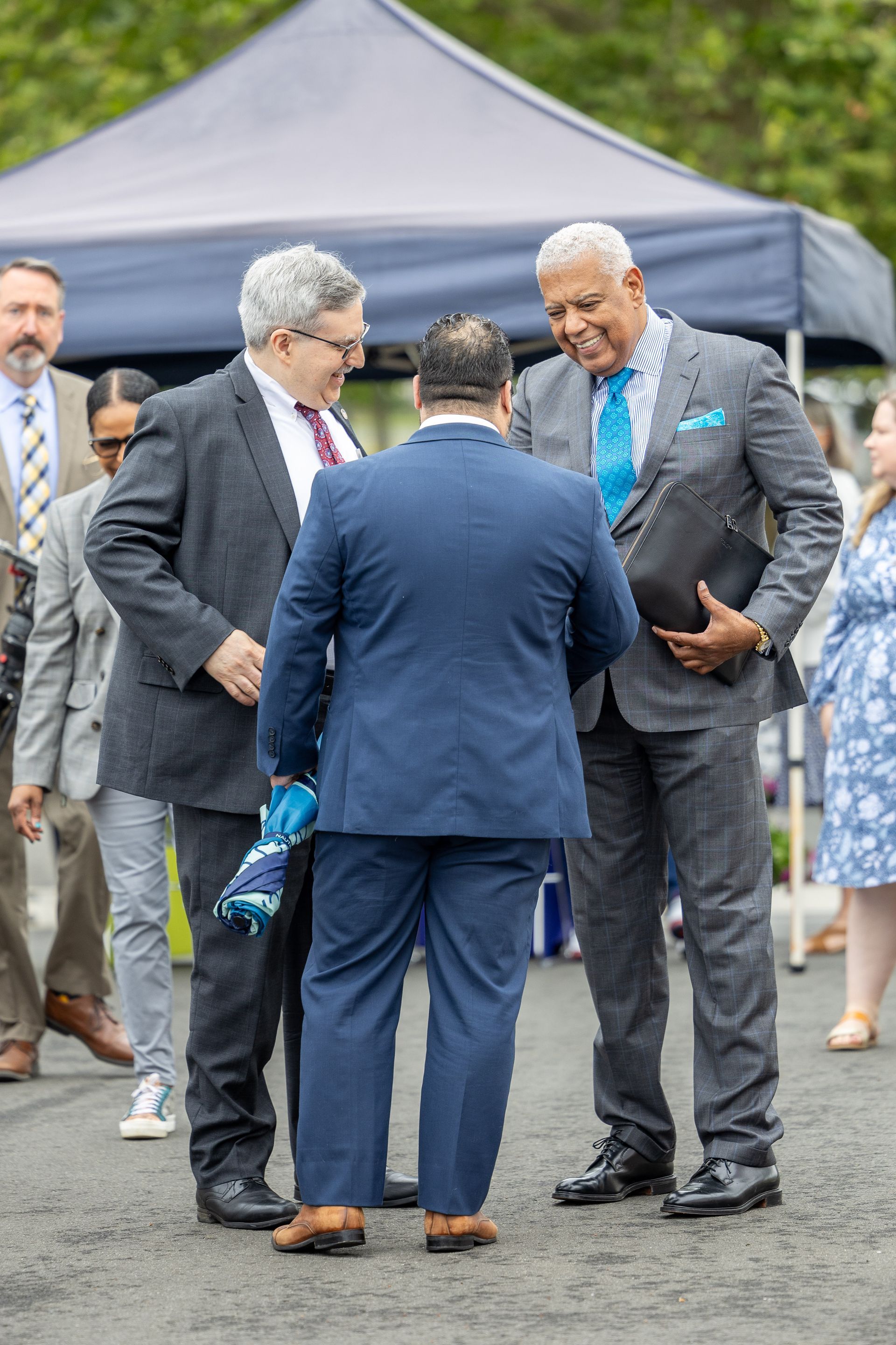 Three men in suits chatting outdoors at a formal event under a canopy.