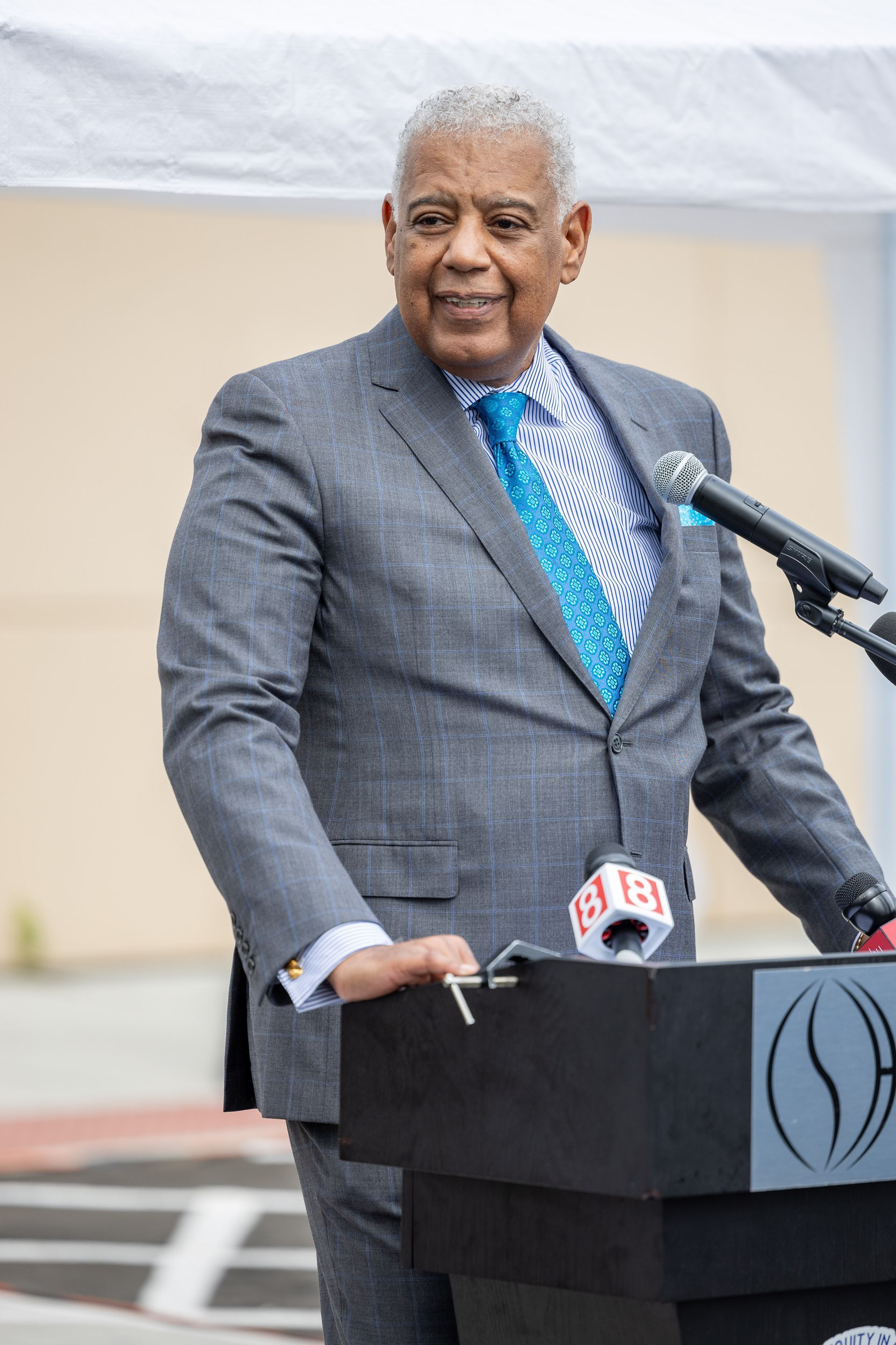 Man in a gray suit speaking at a podium with microphones outdoors