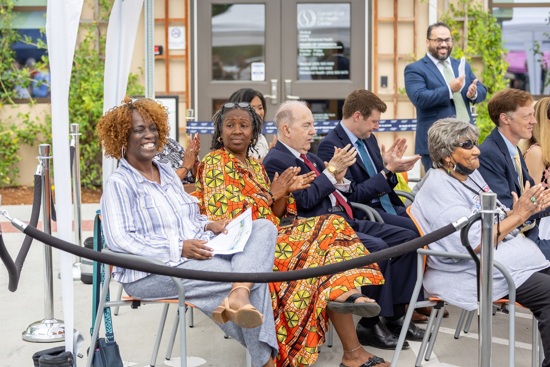 Seated audience applauding at an outdoor event, with people smiling and a man standing nearby.