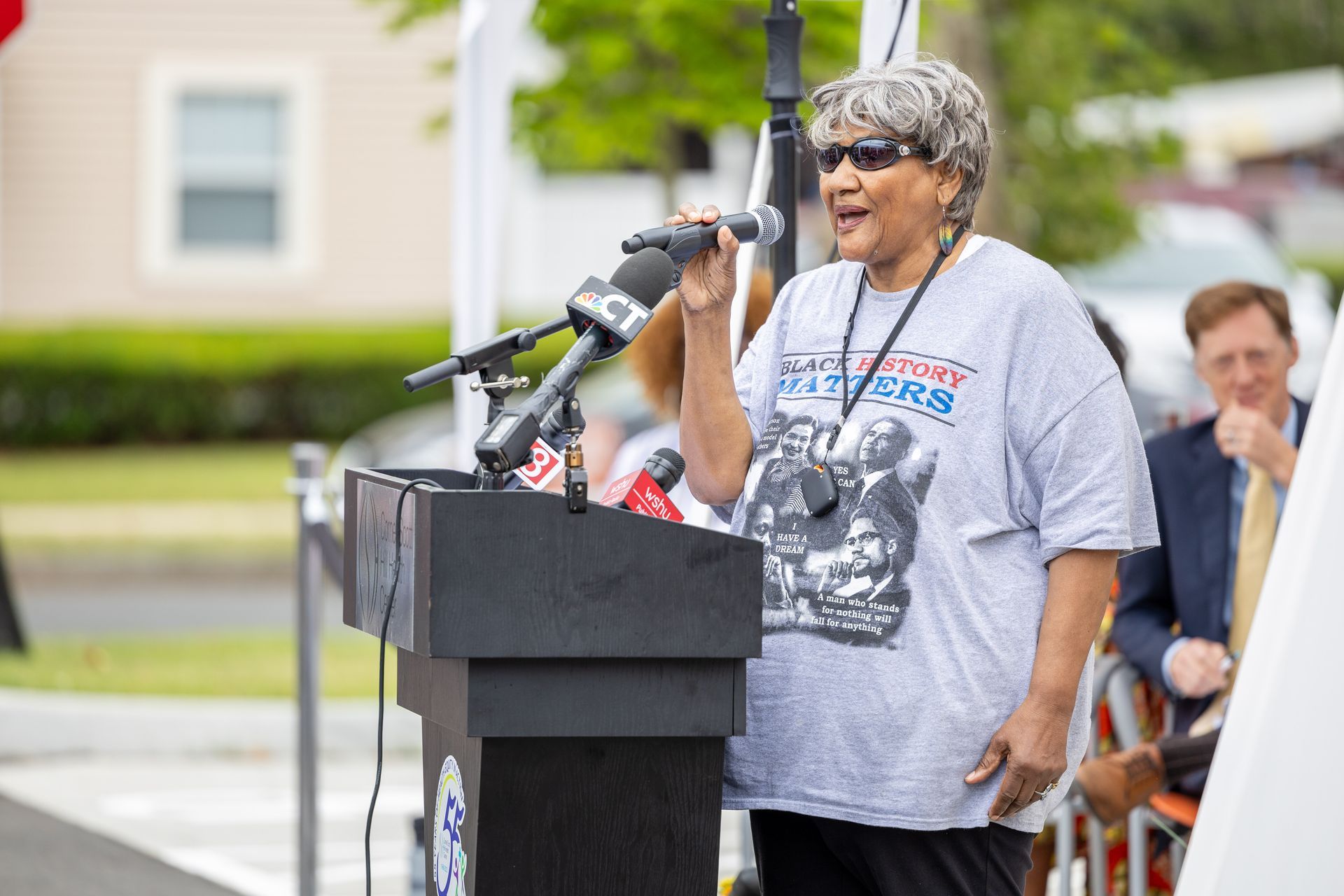 Speaker addressing crowd at outdoor podium with microphones, wearing a gray T-shirt and cap