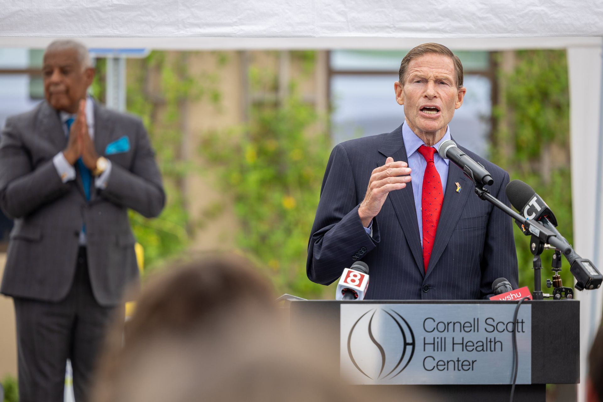 Speaker at podium during outdoor event, with another man standing behind him and sign for Cornell Scott Hill Health Center.