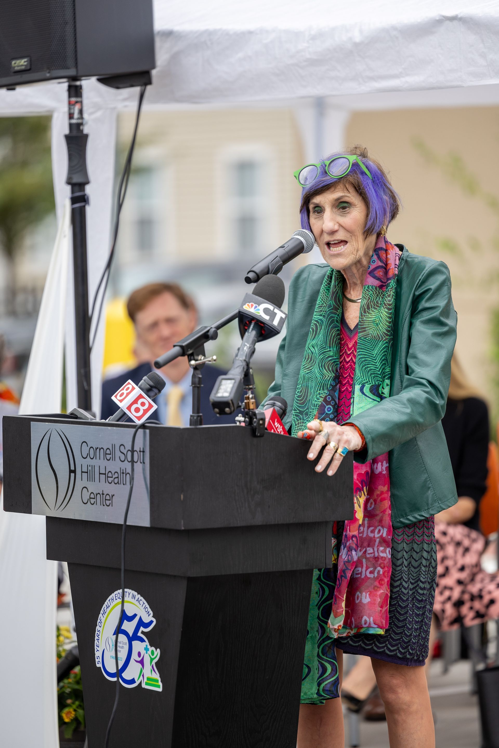 Woman speaking at a podium outdoors, wearing a green jacket and colorful scarf, with microphones and a tent overhead