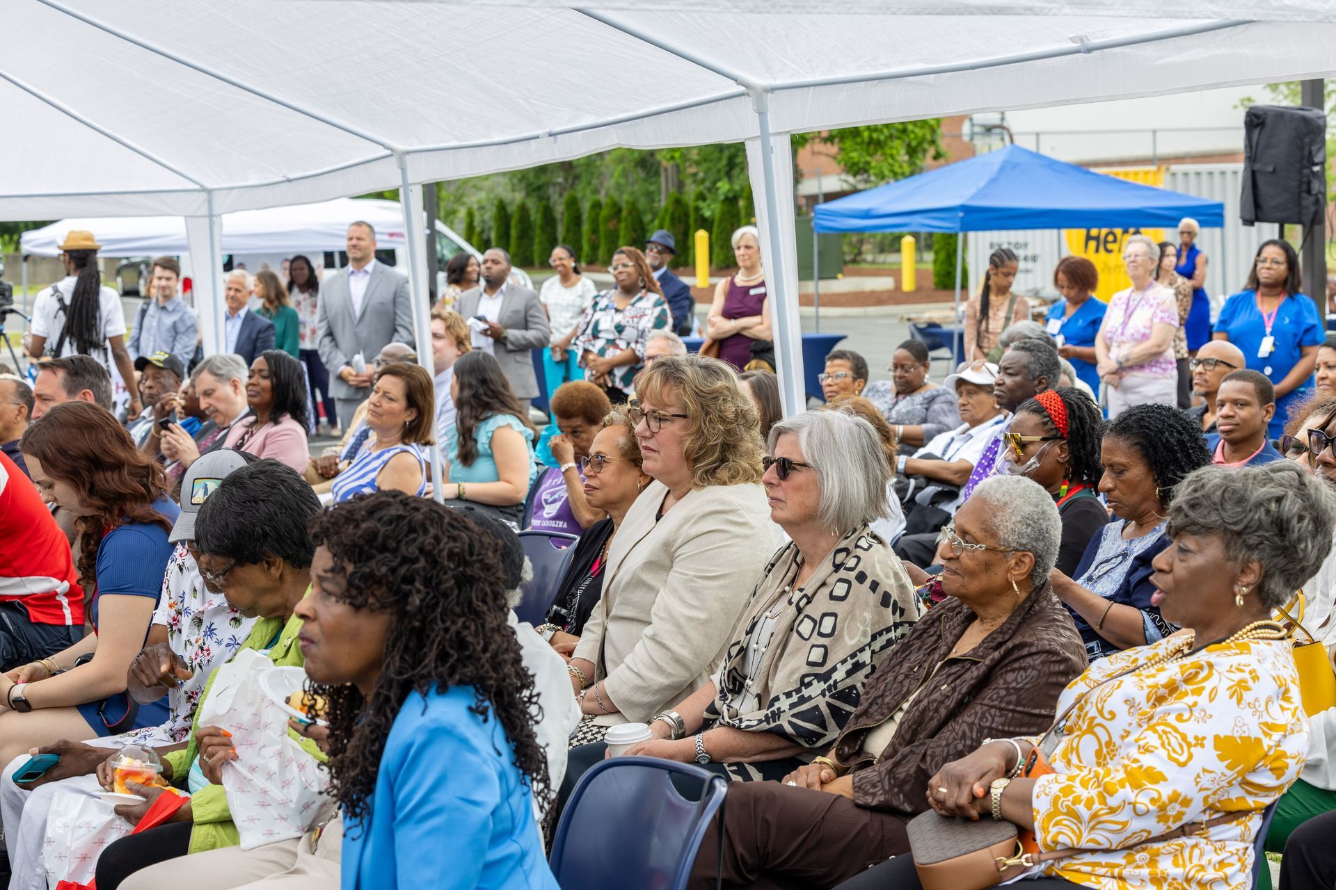 Audience seated under a white tent at an outdoor community event, with a speaker visible in the background
